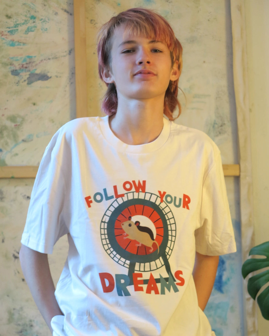 Person trägt Illustration eines Hamsters im Laufrad mit der Typografie ‚Follow Your Dreams‘ im collageartigen Cartoon-Stil auf einem weißen T-Shirt