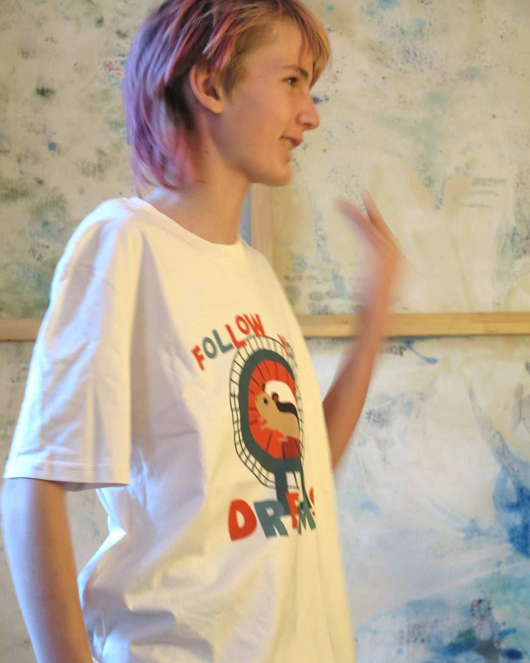 winkende Person trägt Illustration eines Hamsters im Laufrad mit der Typografie ‚Follow Your Dreams‘ im collageartigen Cartoon-Stil auf einem weißen T-Shirt, Seitenansicht