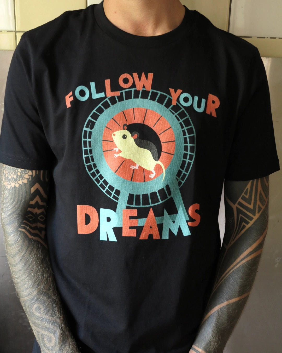 Person trägt Illustration eines Hamsters im Laufrad mit der Typografie ‚Follow Your Dreams‘ im collageartigen Cartoon-Stil auf einem schwarzen T-Shirt aus Baumwolle