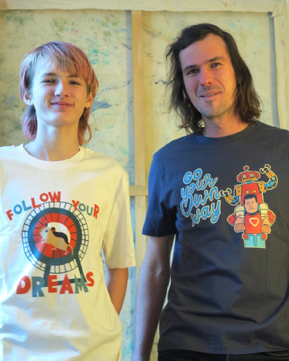 eine junge Person trägt ein weißes T-Shirt mit der Aufschrift 'Follow Your Dreams', welches einen Hamster im Laufrad abbildet. Sie steht neben einer Person mit der T-Shirt-Aufschrift 'Go your own Way'