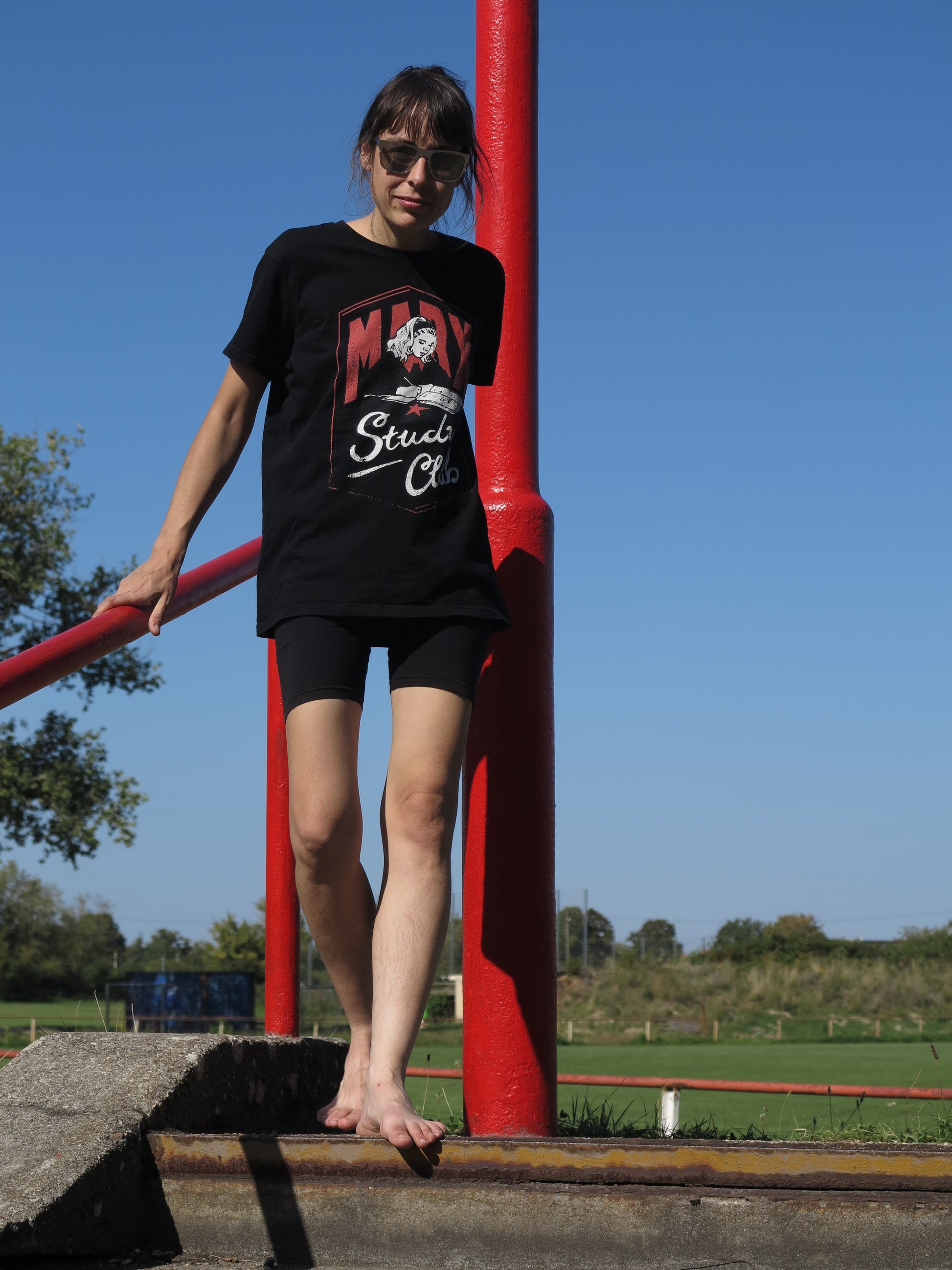 Barfüßige Person trägt Sonnenbrille und T-Shirt mit Retro-Illustration eines Mädchens über einem Buch mit ‚Marx Study Club‘ Typografie, Bezug zu Karl Marx und Kommunismus